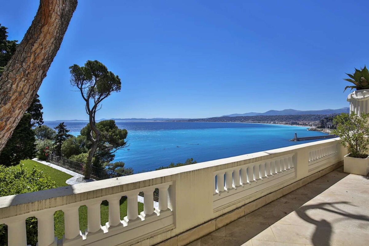 A stunning panoramic view from a balcony overlooking a vibrant turquoise sea, distant coastline, and city, all under a clear blue sky.