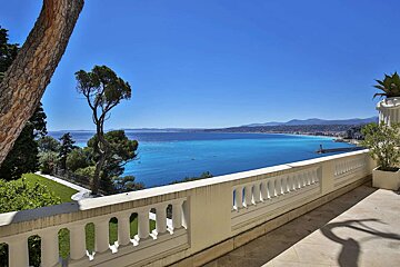 Sunny balcony overlooking a stunning vivid blue sea, distant city, and clear sky, framed by trees and a white balustrade.
