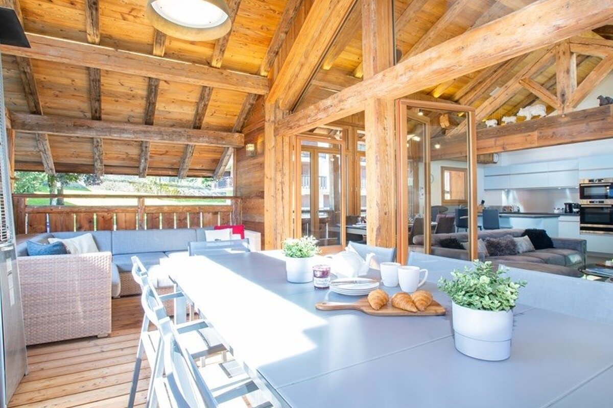 Bright rustic wooden chalet with a long dining table set with pastries, an outdoor patio lounge, and high beamed ceilings.