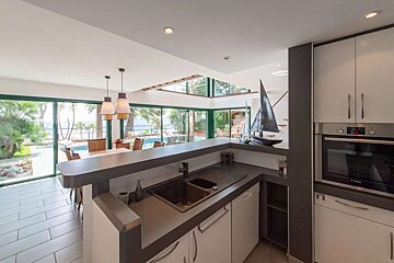 A sleek kitchen with dark counters and white cabinets overlooks an open-plan dining area, a pool, lush greenery, and the ocean beyond.