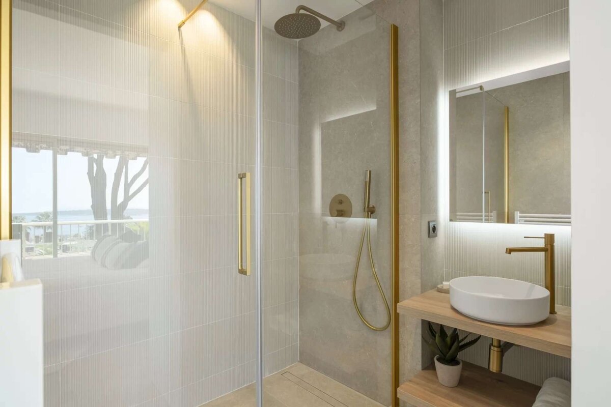 Modern bathroom with a glass shower and gold fixtures. A vanity with a white sink and lighted mirror complements the minimalist design. Trees and water are visible outside.