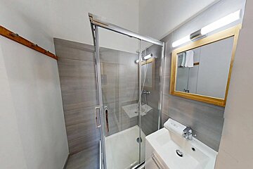 A modern bathroom featuring a glass shower stall, a white vanity with a sink, and a wooden-framed mirror. Grey tiled walls and a wooden wall rack are also visible.