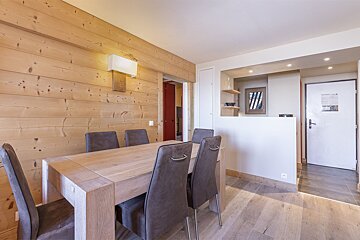 A modern dining area features a light wood table with grey chairs, set against a wood-paneled wall. A hallway with a white door is visible on the right.