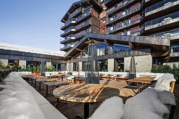 A cozy outdoor patio at a modern ski resort, with wooden tables, white fur-covered seating, and heaters, set against a multi-story hotel.