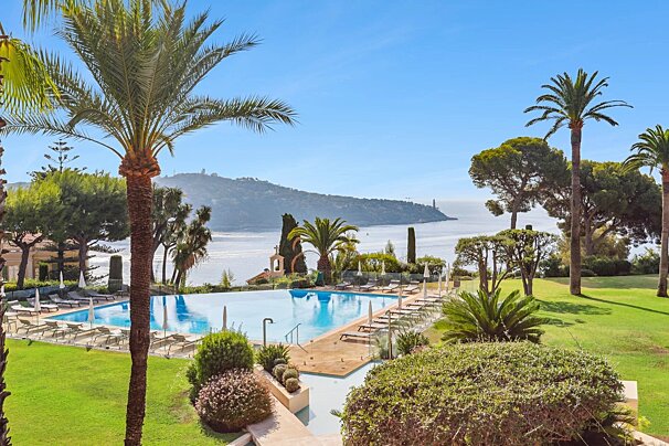 A luxurious infinity pool overlooks a serene bay and distant hills, framed by lush gardens and palm trees under a clear, sunny sky.