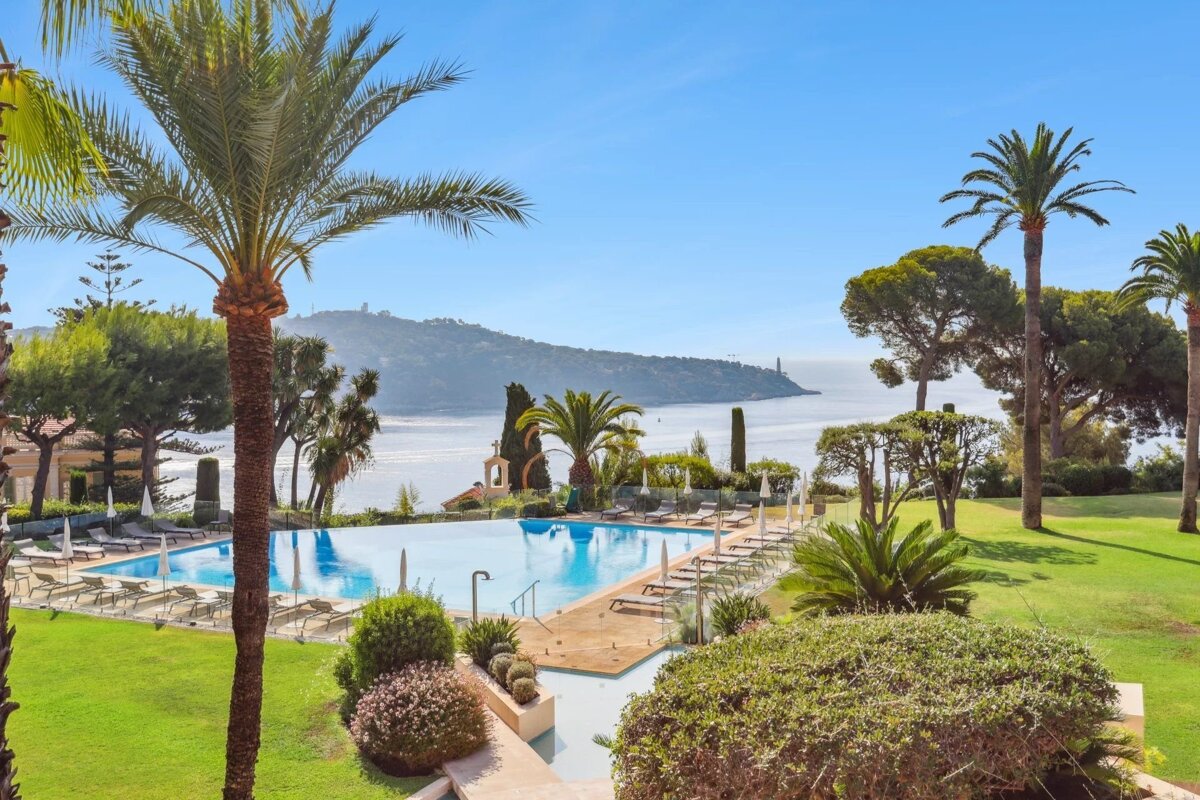 A beautiful infinity pool with lounge chairs and palm trees, overlooking a serene blue bay and distant headland on a sunny day.