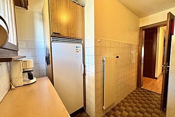 Small kitchen area with a white refrigerator, wood cabinet, coffee maker, beige tiled walls, and a checkered floor leading to a hallway.
