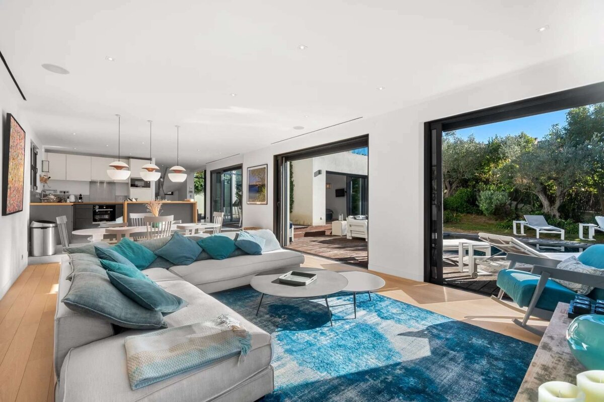 Modern open-plan living area with an L-shaped sofa, blue rug, and kitchen. Large sliding doors offer access to an outdoor patio and lush garden.