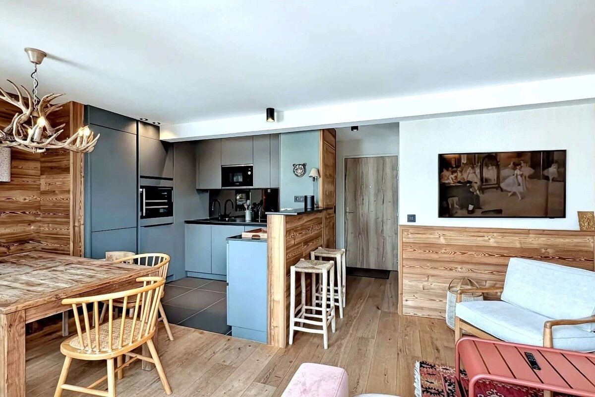 A modern open-plan living space featuring a blue kitchen, rustic wood dining area with antler chandelier, and a TV. Warm wooden accents and floors throughout.