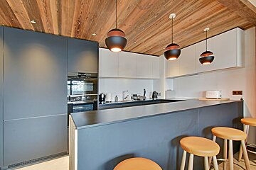A modern kitchen with a wooden ceiling, sleek gray and white cabinets, an island with orange bar stools, and unique black pendant lights.