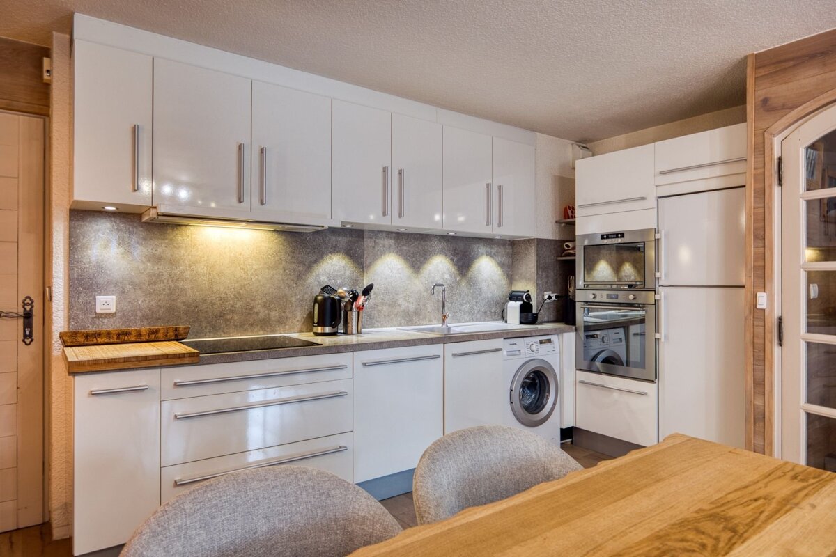 A sleek, modern kitchen with white glossy cabinets, a textured grey backsplash, integrated appliances (washer, oven, fridge), and warm wooden details.