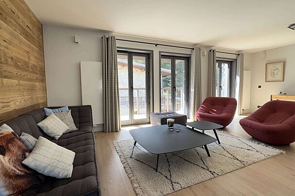 Modern living room with a wooden feature wall, cozy sofa, two red lounge chairs, geometric rug, coffee tables, and bright patio doors.