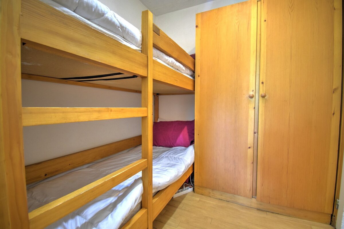 A compact room features light wooden bunk beds with white bedding and a matching wardrobe. A magenta pillow brightens the lower bunk.