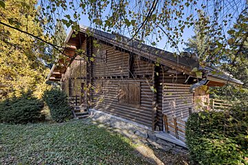 A rustic wooden log cabin is nestled among trees with colorful autumn leaves and green shrubs under a clear blue sky.