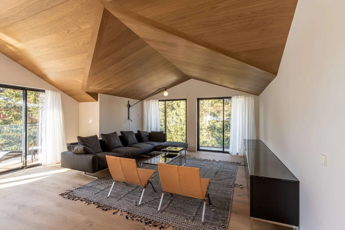 A modern living room features a striking angled wood-paneled ceiling, large windows with tree views, a dark sectional sofa, and light wood floors.