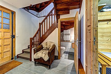 Rustic entry with a prominent wooden staircase, a brown armchair with a fur throw, and an open door revealing a toilet under the stairs. Tiled floor.