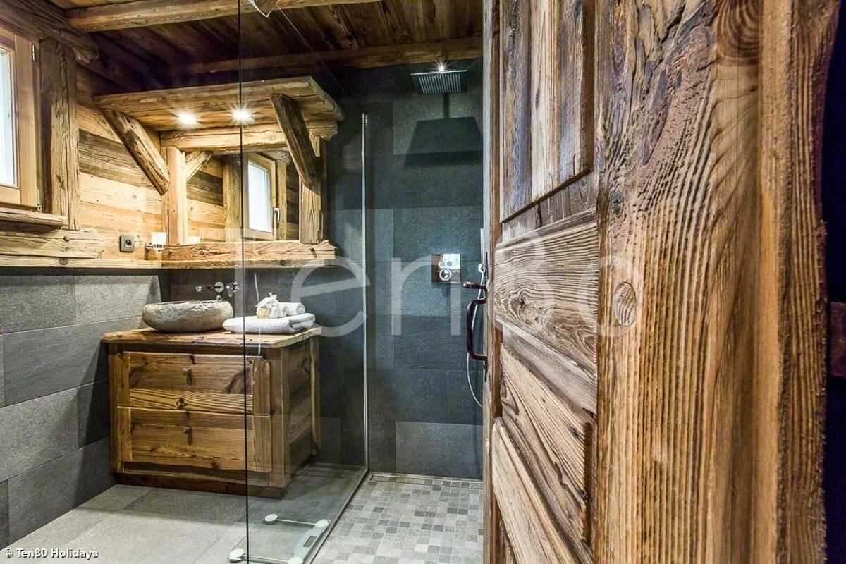 Rustic wooden bathroom with dark grey tiles, a stone sink, a glass-enclosed shower, and a textured wood door in the foreground.