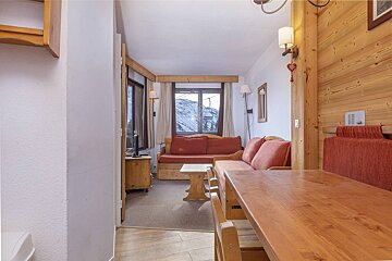 A cozy, rustic living room with wooden furniture, red sofas, and a window view of snowy mountains and a ski lift.