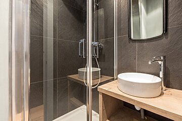 Modern bathroom with glass shower, white vessel sink on a wooden vanity, dark gray tiled walls, and a mirror.
