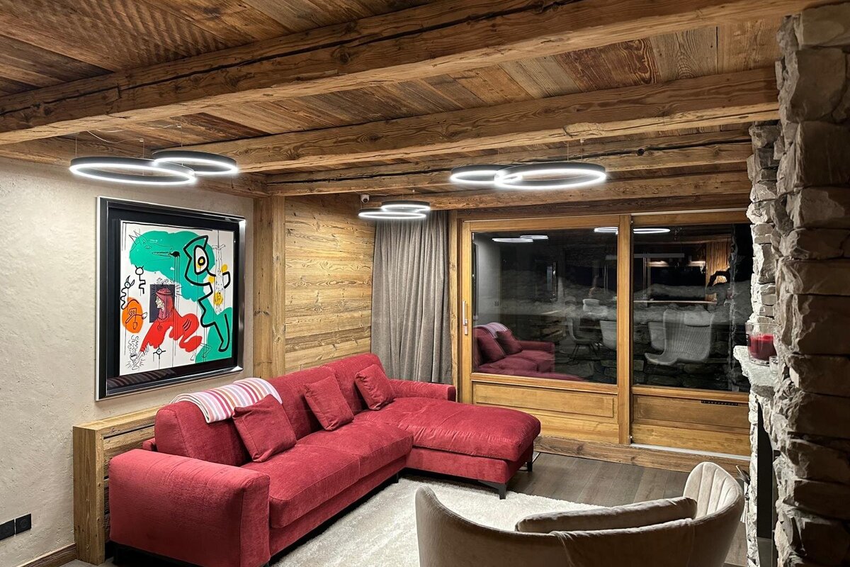 A cozy living room with rustic wooden ceiling, modern ring lights, a red sectional sofa, colorful abstract art, and a large glass door reflecting outside.