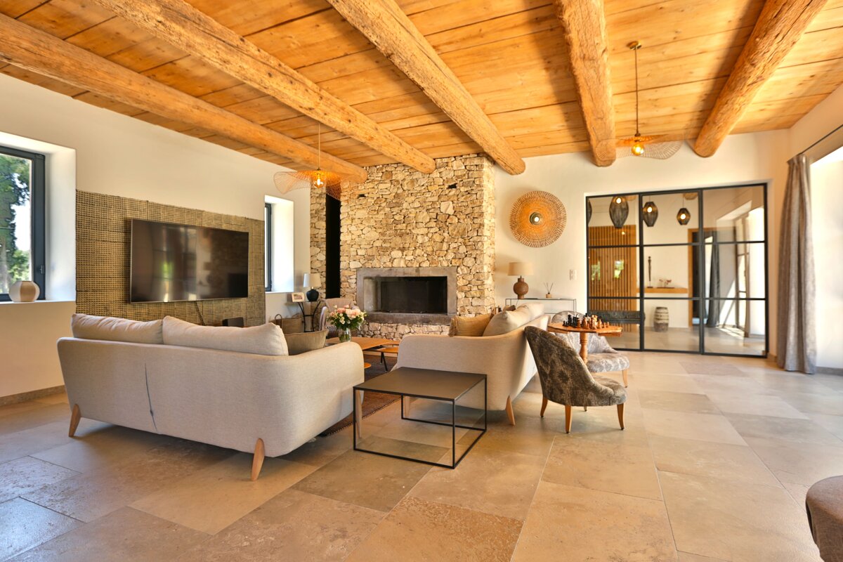A spacious living room blends rustic wood beams and a stone fireplace with modern light sofas, tiled floor, and a sleek black-framed glass partition.