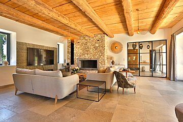 A spacious living room blends rustic wood beams and a stone fireplace with modern light sofas, tiled floor, and a sleek black-framed glass partition.