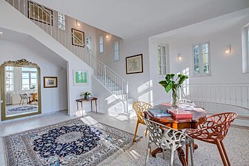 Sunlit, multi-level entryway with a white staircase, ornate mirror, patterned rug, and a dining table with colorful chairs.