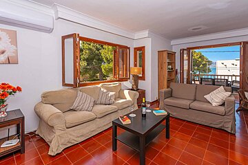 A cozy living room with reddish-brown tiled floors, two couches, and windows offering views of green trees and the blue sea.
