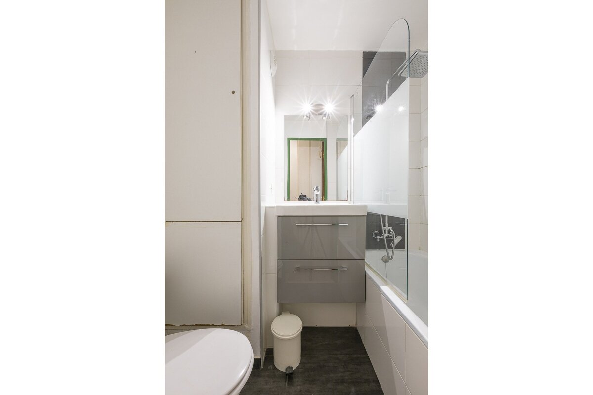 Compact modern bathroom with white and gray decor. Features a toilet, sink vanity, and bathtub with a glass shower screen.