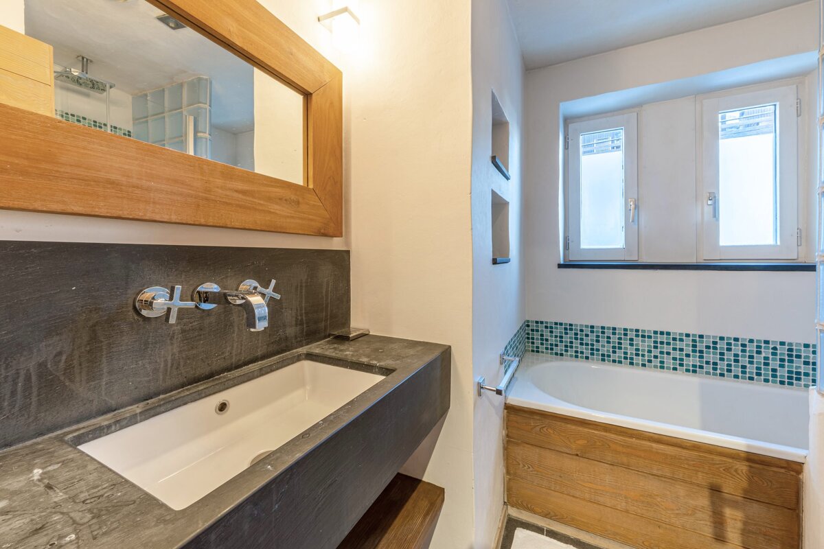 Bathroom with wooden mirror, stone sink, wood-paneled bathtub, green mosaic tile trim, and two windows.