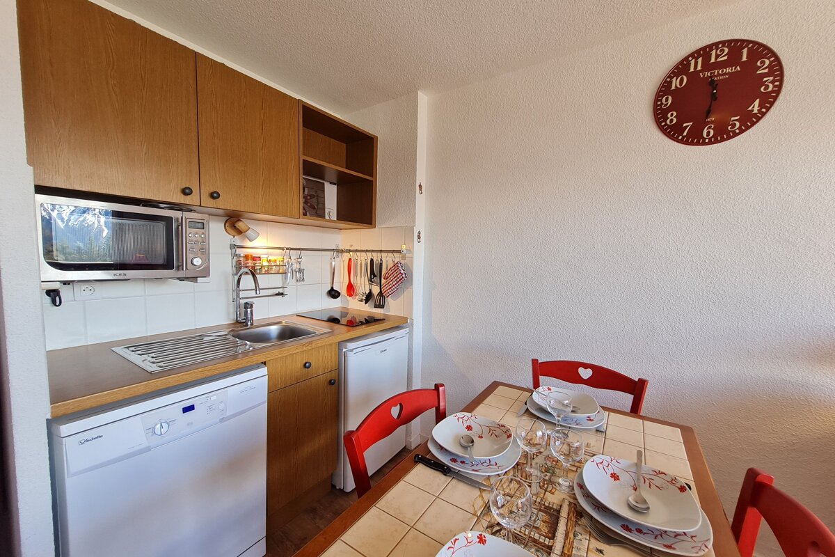 A compact kitchenette with wood cabinets and white appliances next to a dining table set with dishes and red chairs featuring heart cutouts. A red clock hangs on the wall.