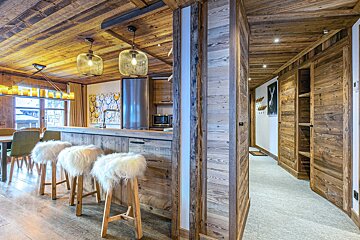 A cozy, rustic-modern chalet interior featuring extensive wood paneling, a kitchen island with fuzzy stools, and a hallway.