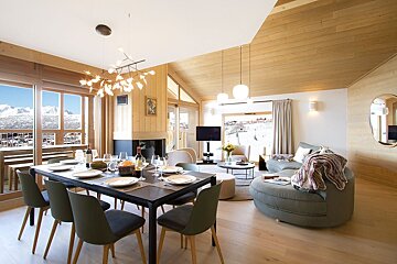 A modern chalet living/dining room featuring extensive wood, a set table, and plush seating. Large windows reveal breathtaking snowy mountain views.