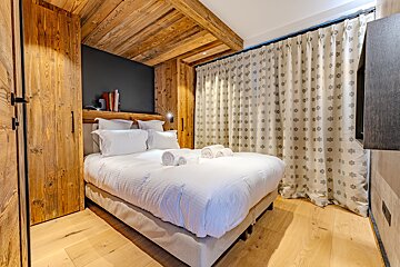 Cozy alpine-style bedroom featuring rustic wood paneling on the walls and ceiling, a neat double bed, and patterned curtains.