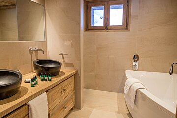 Modern rustic bathroom with twin black stone sinks on a wooden vanity, a white tub, and a window revealing snowy views.