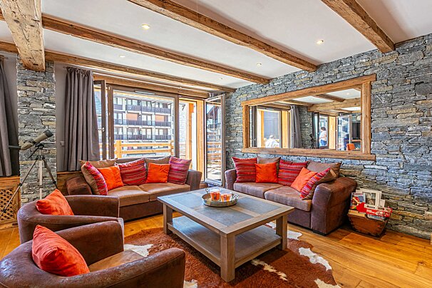 A rustic, cozy living room with stone walls, wooden beams, brown sofas with orange cushions, and a large window balcony view.