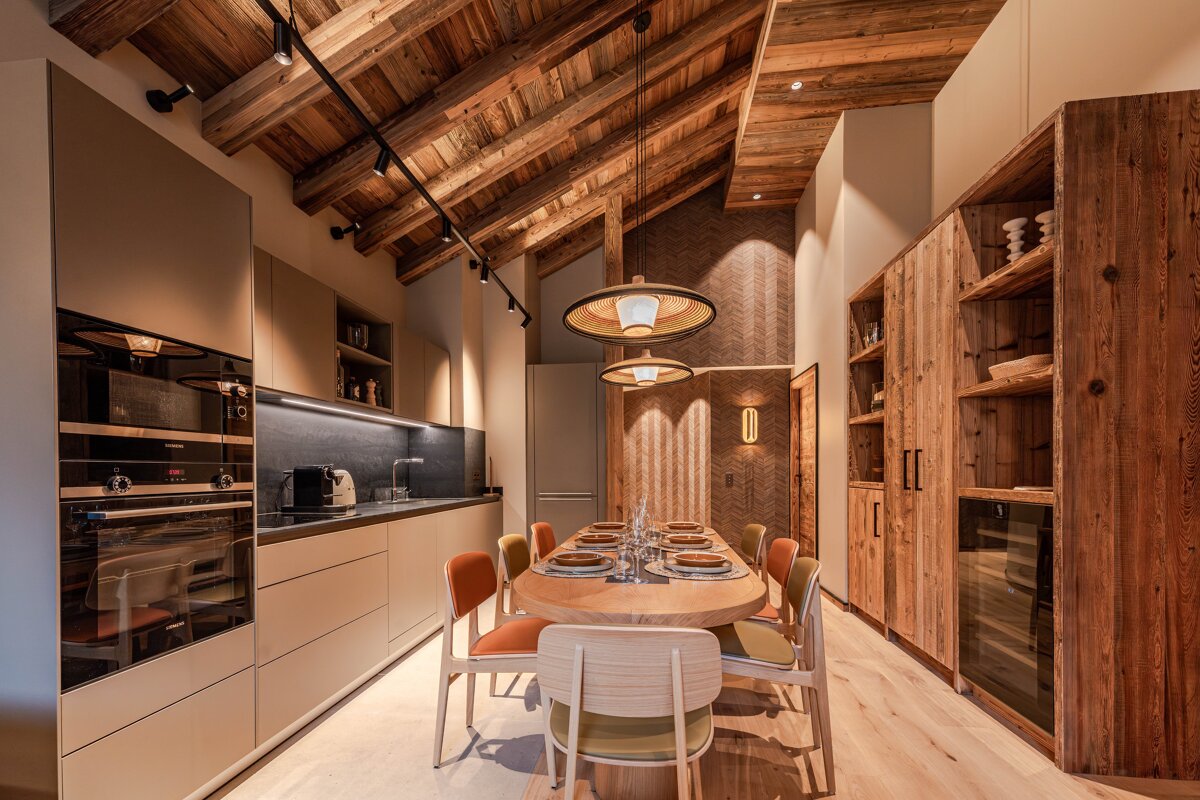A warm, modern chalet dining area and kitchen with an exposed wooden ceiling, sleek cabinets, and an elegant dining table.