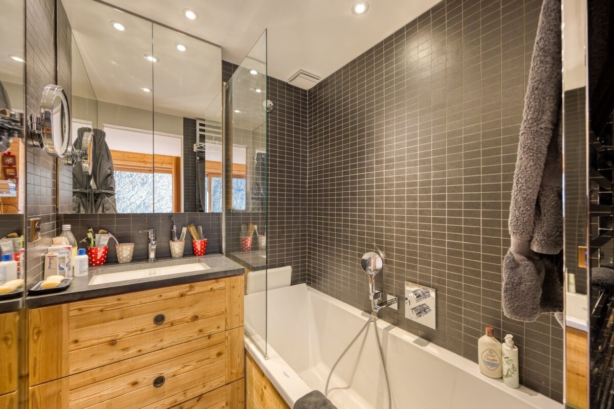 A modern bathroom features dark square tiles, a rustic wooden vanity with a sink, a bathtub with a glass screen, and a large mirrored cabinet.