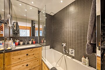 A modern bathroom features dark square tiles, a rustic wooden vanity with a sink, a bathtub with a glass screen, and a large mirrored cabinet.