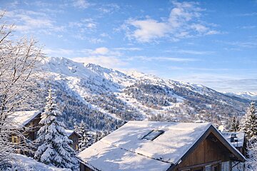 Snow-covered wooden chalets nestled in an alpine forest with a majestic, snow-capped mountain under a bright blue sky.