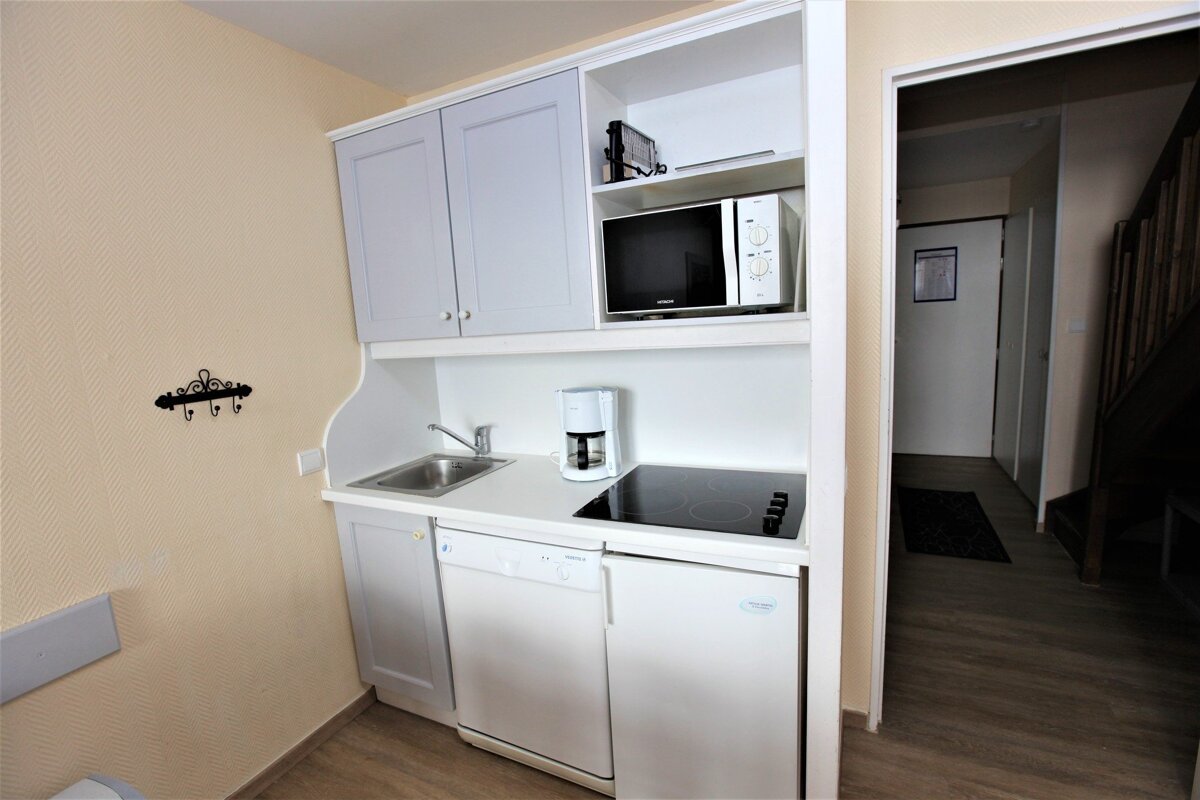 A compact, modern kitchenette with light grey cabinets, sink, microwave, stovetop, coffee maker, and appliances. A hallway and staircase are visible.