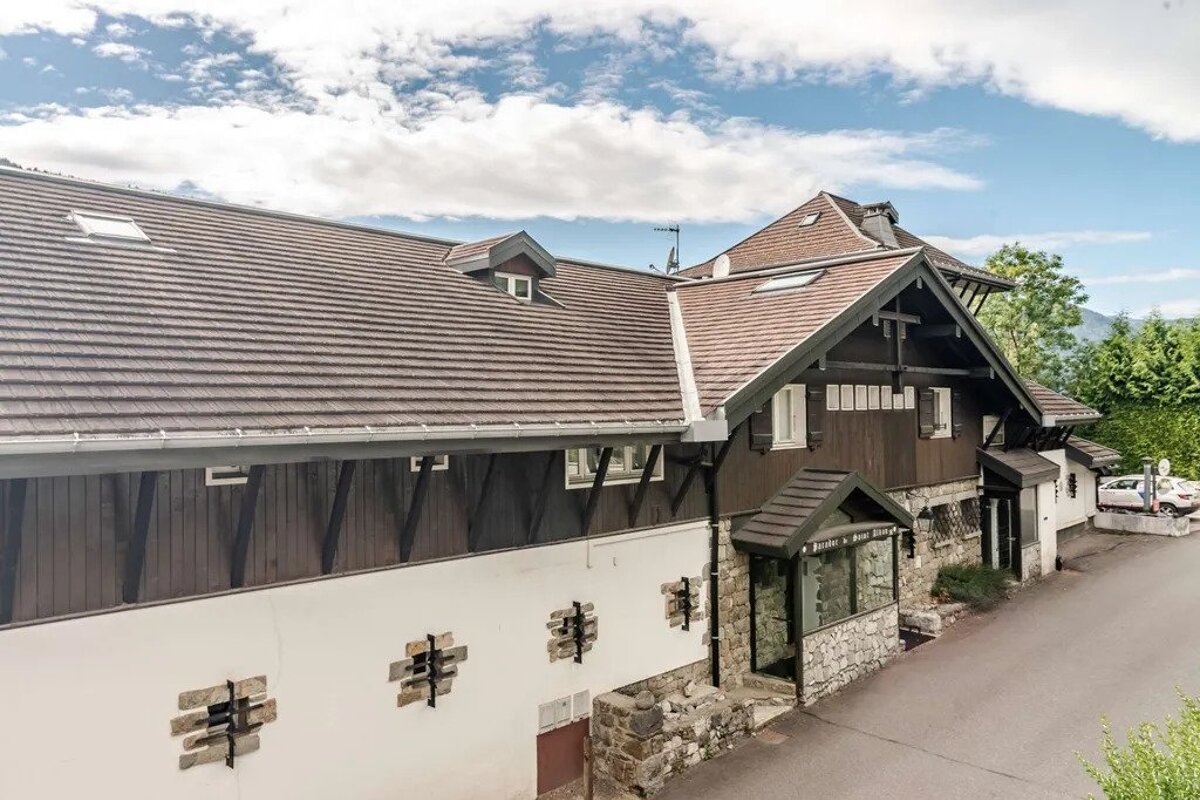 A rustic multi-story building with a brown tiled roof, dark wooden upper levels, and white lower walls with stone accents. Mountains and trees are in the background under a cloudy sky.