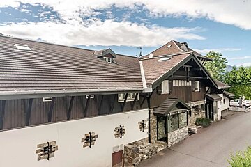 A chalet-style building with brown roof tiles and a dark wood/white stone facade stands by a road, with mountains and trees in the background under a cloudy sky.