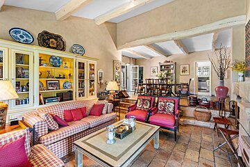 Cozy, eclectic living room with plaid sofa, red chairs, tile floor, beamed ceiling, and bookshelves. Dining area visible behind.