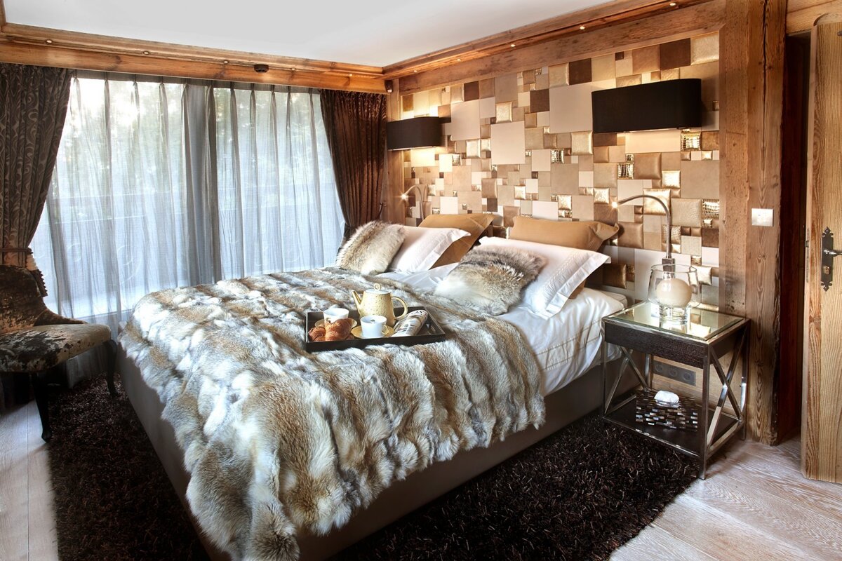 A chic bedroom with a large fur-covered bed, breakfast tray, striking tiled headboard, and warm wooden details.