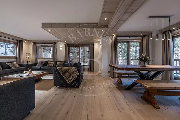 Spacious, modern chalet-style living/dining room with light wood floors, gray sofas, a rustic wooden dining table, and large windows with a snowy view.