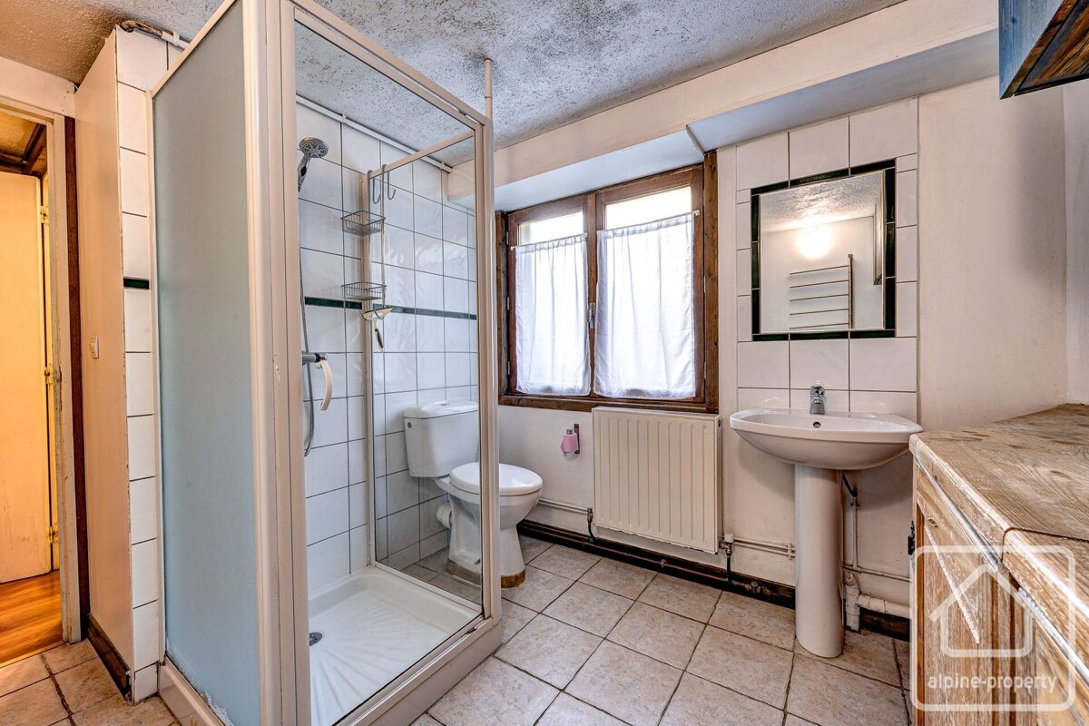 A small bathroom with a glass shower, toilet, pedestal sink, mirror, and a window with white curtains. It has tiled floors and walls.