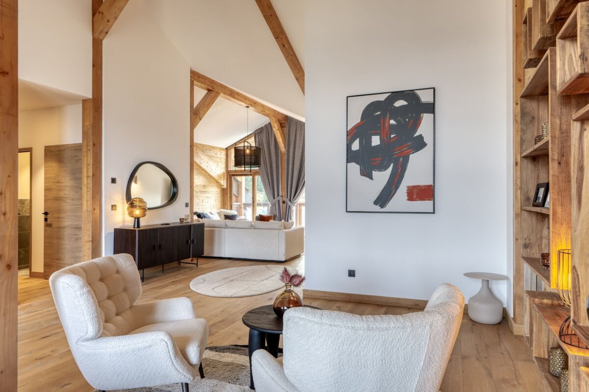 Stylish living room with white armchairs, abstract art, wooden beams, and a glimpse of a modern lounge area.