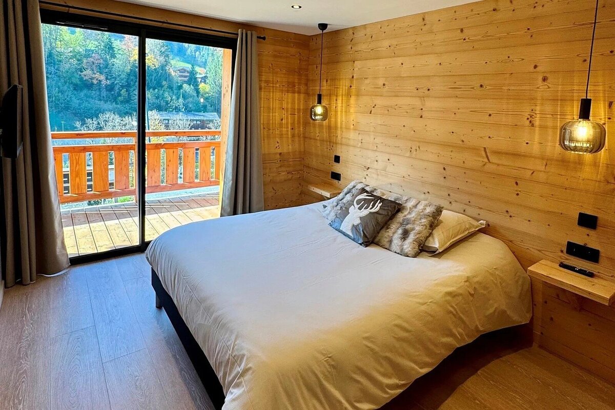 A cozy, wood-paneled bedroom features a large bed with a deer-themed pillow and a glass door opening to a balcony with a scenic mountain view.