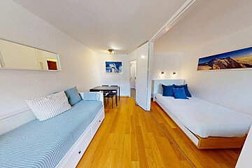 Bright studio apartment with wooden floors, a blue daybed, a single bed, and a small dining table in the background.